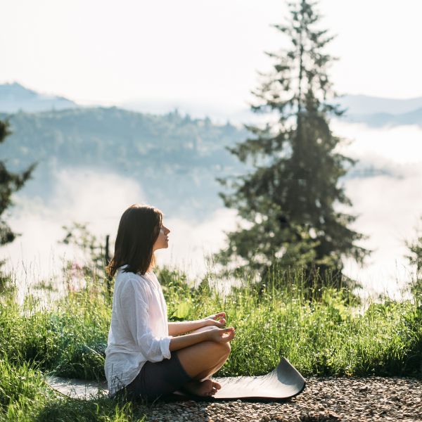 Woman meditating outdoors on a yoga mat, surrounded by nature and morning mist, embodying mindfulness, relaxation, and inner balance. A representation of stress relief, emotional well-being, and self-care—core elements of wellness coaching at Renew Strength and Wellness.